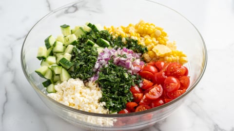 A glass bowl filled with chopped cucumber, cherry tomatoes, corn salad, red onion, fresh herbs, and crumbled feta cheese, all beautifully arranged in separate sections on a white marble surface.