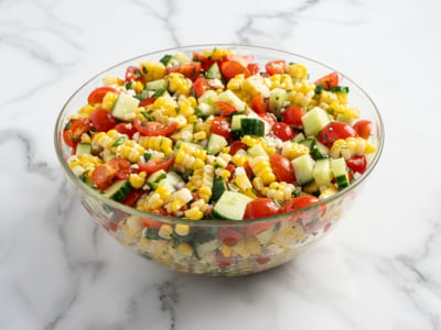 A glass bowl filled with a vibrant Corn Salad made of cherry tomatoes, cucumber, and red onion sits on a white marble surface.