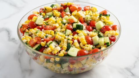A glass bowl filled with a vibrant Corn Salad made of cherry tomatoes, cucumber, and red onion sits on a white marble surface.