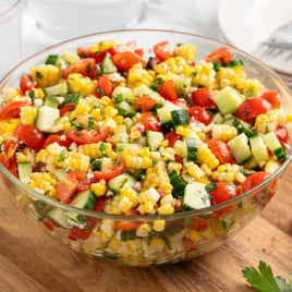 A glass bowl filled with vibrant Corn Saladโcorn, cherry tomatoes, cucumbers, red onion, and fresh herbsโsits on a wooden surface. Plates, forks, and a sprig of parsley are visible nearby.