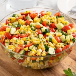 A glass bowl filled with vibrant Corn Salad made of cherry tomatoes, cucumbers, red onions, and herbs sits on a wooden surface, with plates and utensils in the background.