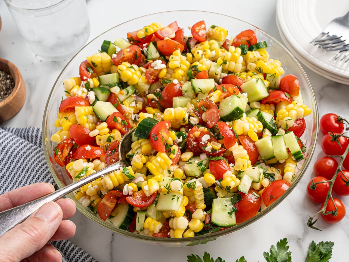 A hand holds a spoon in a glass bowl of vibrant corn salad with cherry tomatoes, cucumber, and herbs on a marble surface, surrounded by a glass of water, pepper, a towel, plates, forks, and vine tomatoes.