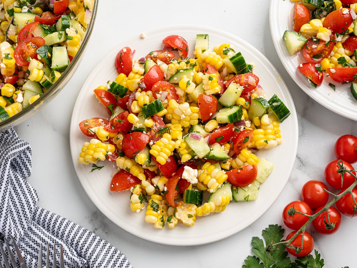 A plate of fresh corn salad with cherry tomatoes, cucumber, herbs, and feta cheese, with more salad in a bowl and a plate nearby. Cherry tomatoes and parsley garnish the marble surface.