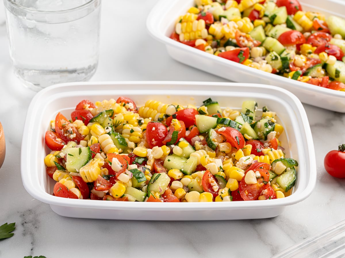 A plastic container filled with vibrant Corn Salad&mdash;featuring corn, cherry tomatoes, cucumber, and herbs&mdash;sits on a white surface next to a glass of water and another fresh salad container.