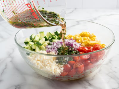 A glass bowl of Corn Salad with chopped cucumber, red onion, cherry tomatoes, corn, and feta cheese is being drizzled with herbed vinaigrette from a measuring cup on a marble countertop.