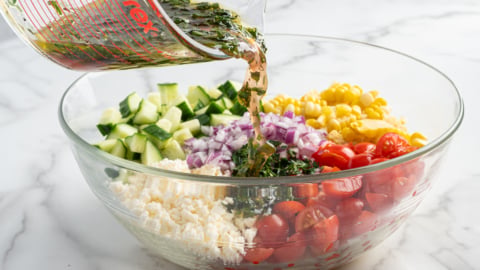 A glass bowl of Corn Salad with chopped cucumber, red onion, cherry tomatoes, corn, and feta cheese is being drizzled with herbed vinaigrette from a measuring cup on a marble countertop.
