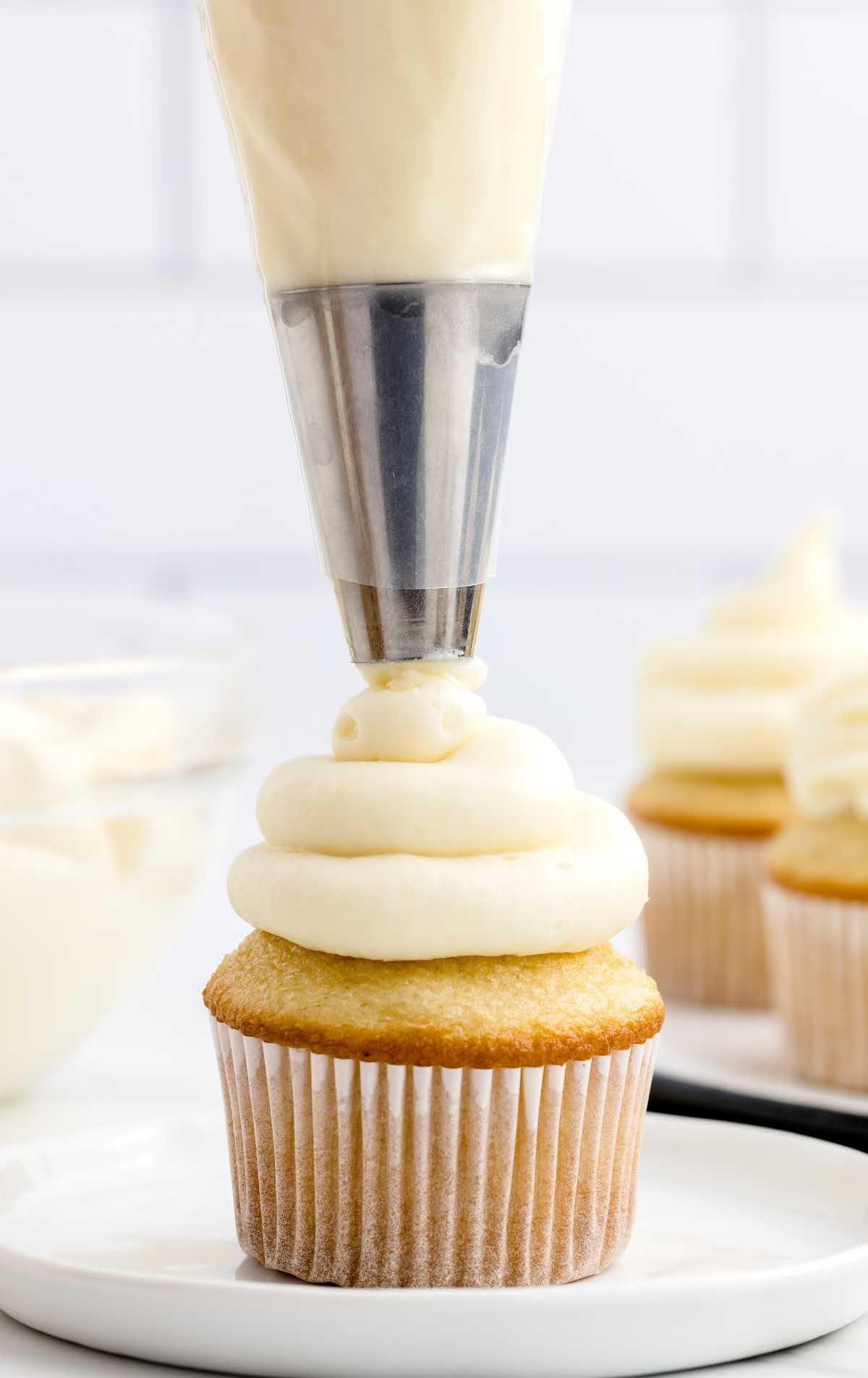 a close up of Cream Cheese Frosting on a cupcake and on a plate with a piping bag
