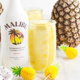 a tall container of Pineapple Spears in Malibu Rum with a bottle of Malibu and pineapples