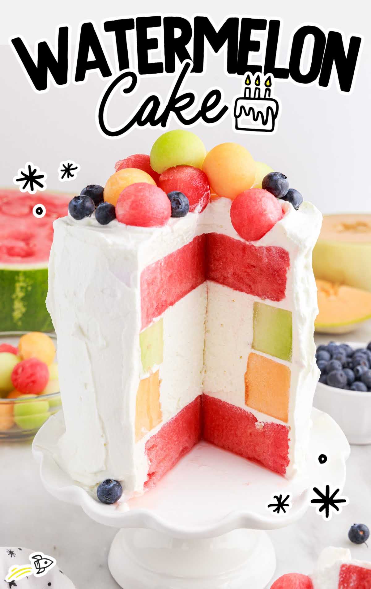 Watermelon Cake on a cake stand with a slice taken out topped with melon balls and blueberries