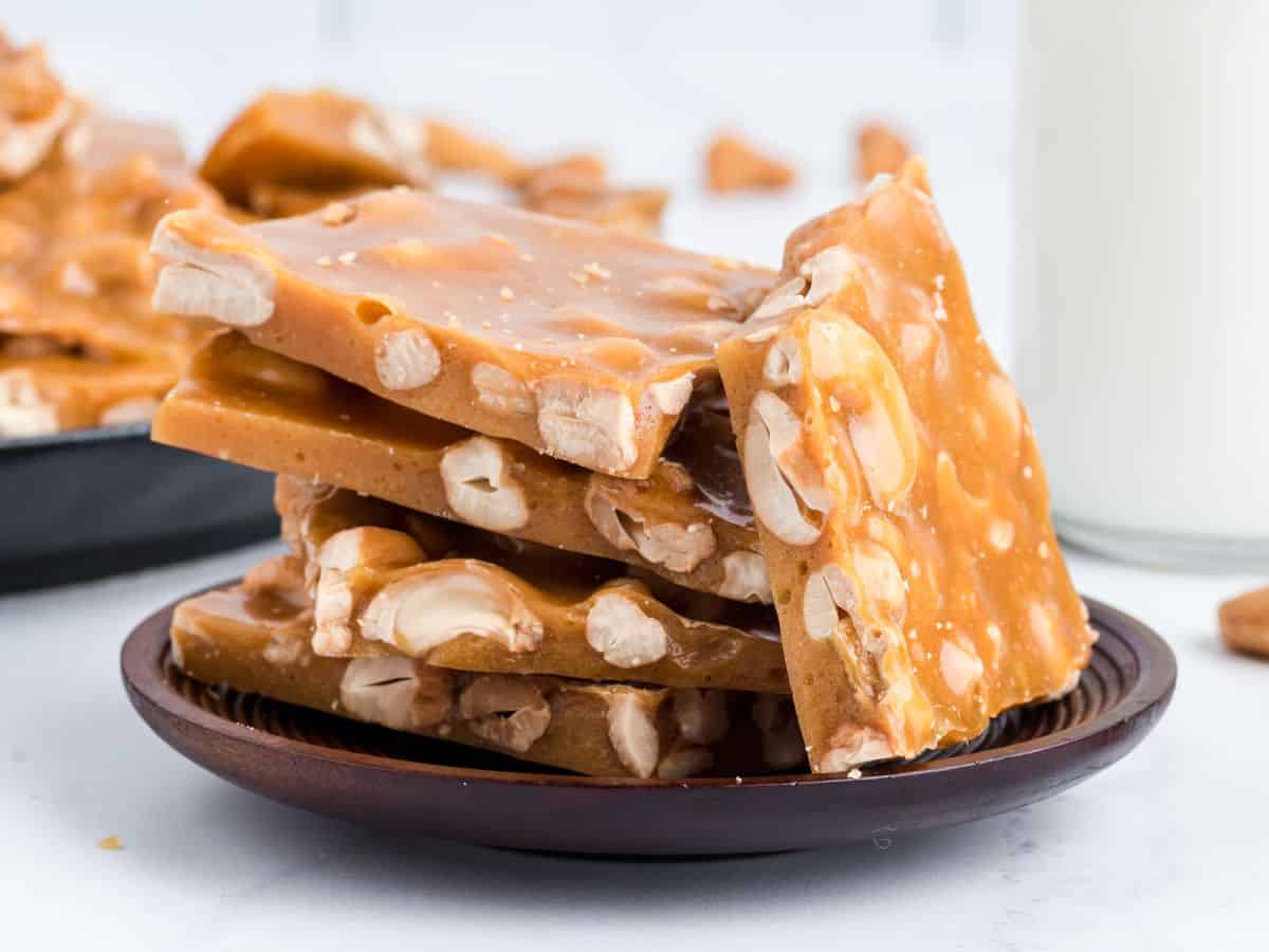 A stack of golden brown cashew brittle pieces on a dark plate, with visible nuts embedded, sits next to a glass of milk on a white surface.