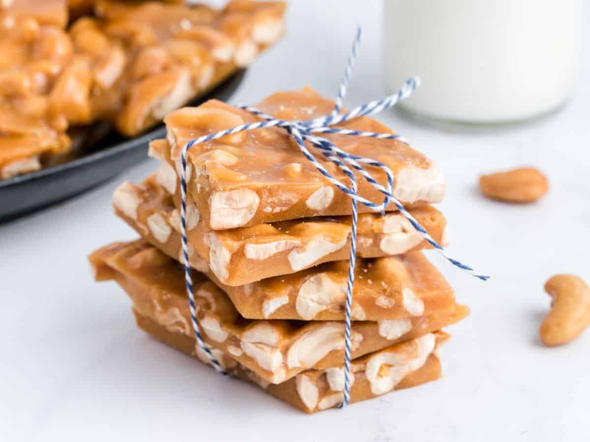 A stack of cashew brittle tied with blue and white string sits on a white surface, with more brittle and a glass of milk in the background.