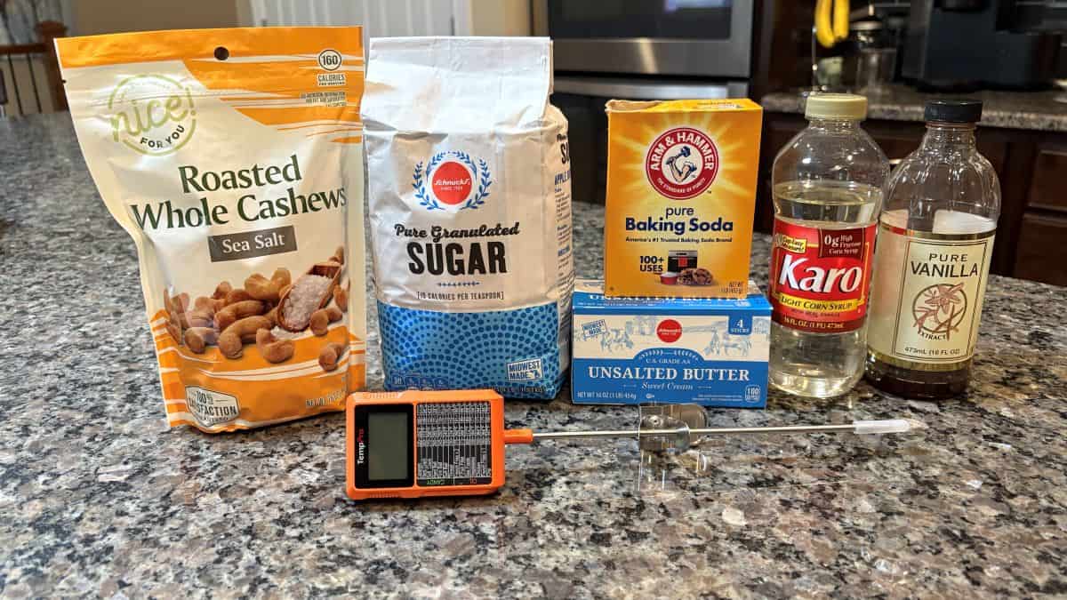 Bags of roasted cashews and sugar, butter, baking soda, corn syrup, vanilla, and a digital thermometer are arranged on a speckled kitchen countertop—everything you need to craft homemade Cashew Brittle.