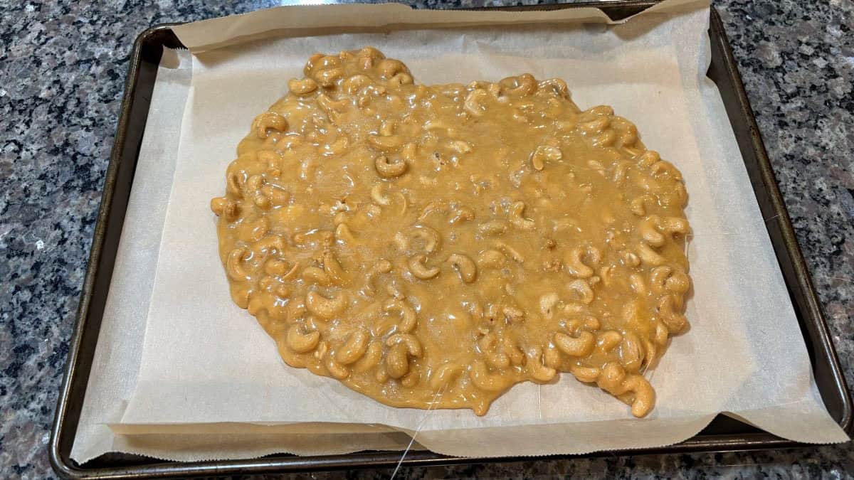A baking sheet lined with parchment paper holds a large, uneven slab of homemade Cashew Brittle, cooling on a granite countertop. The golden surface of the Cashew Brittle glistens invitingly as it hardens to perfection.