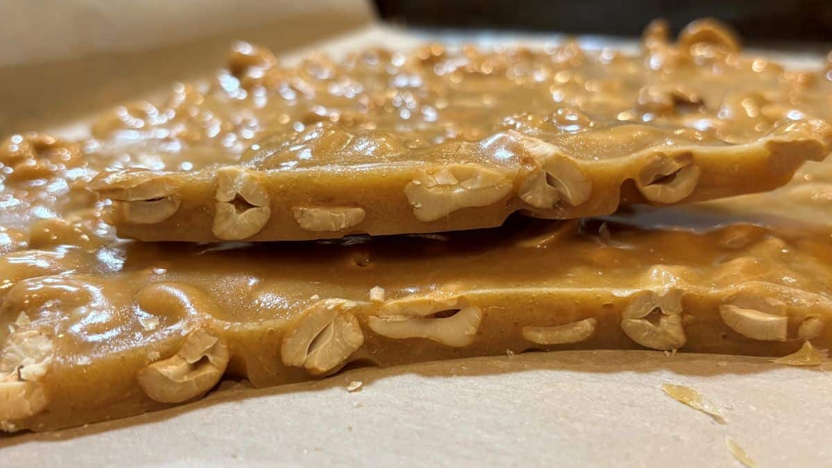 Close-up of two pieces of homemade peanut brittle and cashew brittle stacked on parchment paper, showcasing the glossy, golden-brown candy with embedded peanuts and cashews.