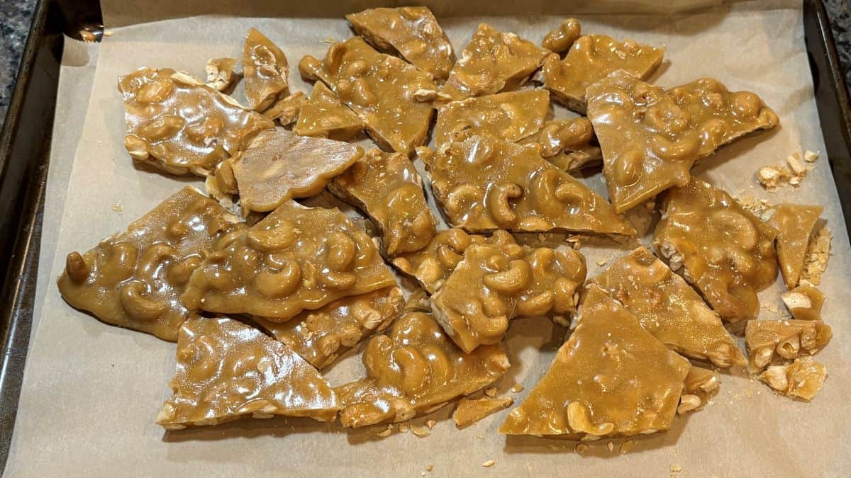 Broken pieces of homemade Cashew Brittle rest on parchment paper atop a baking sheet, their glossy caramelized sugar perfectly coating each cashew nut.