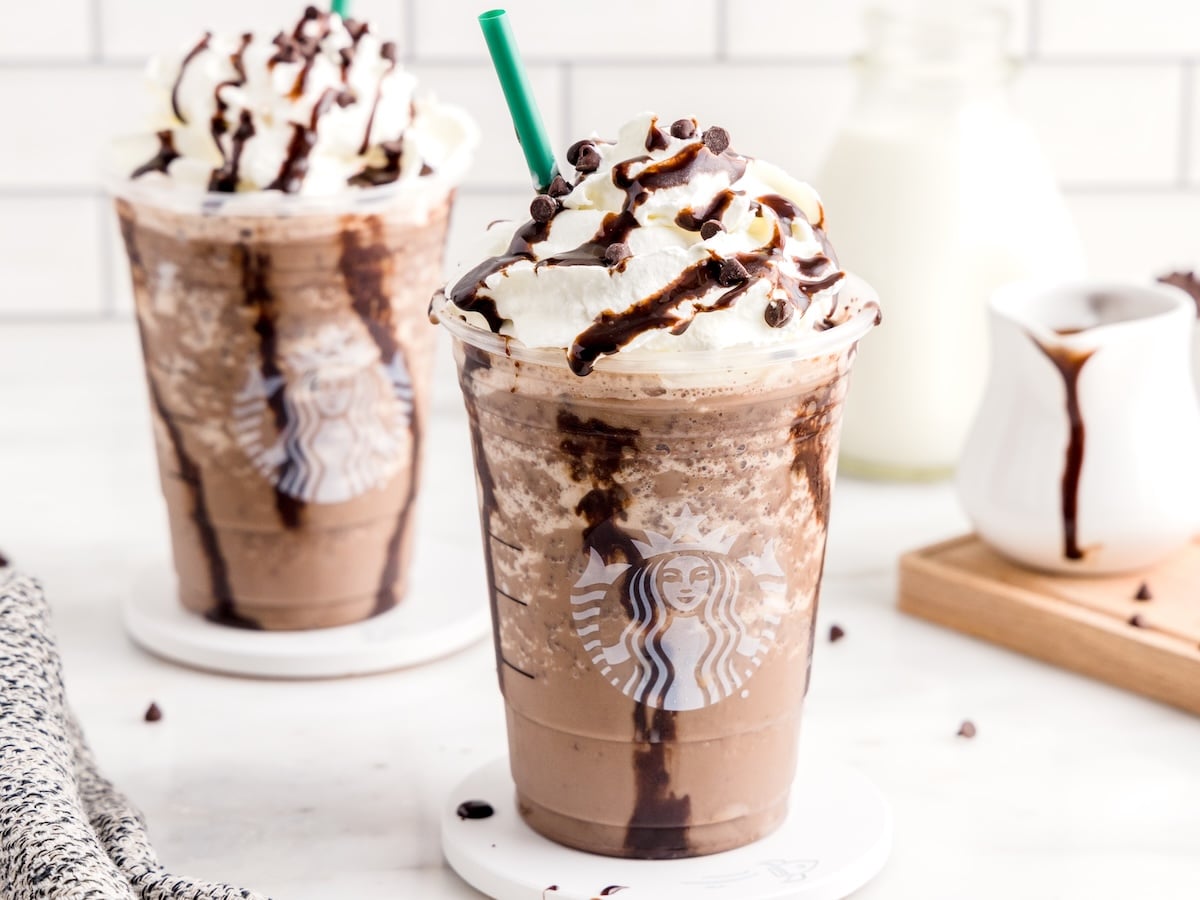 Two Double Chocolate Chip Frappuccinos topped with whipped cream, chocolate drizzle, and chocolate chips sit on white coasters. A green straw is in the front drink, with milk and chocolate sauce in the background.