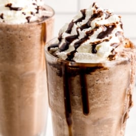 Two tall glasses filled with creamy Double Chocolate Chip Frappuccino milkshakes are topped with whipped cream and drizzled with chocolate syrup. The milkshakes look thick and cold, with chocolate dripping down the glass sides.