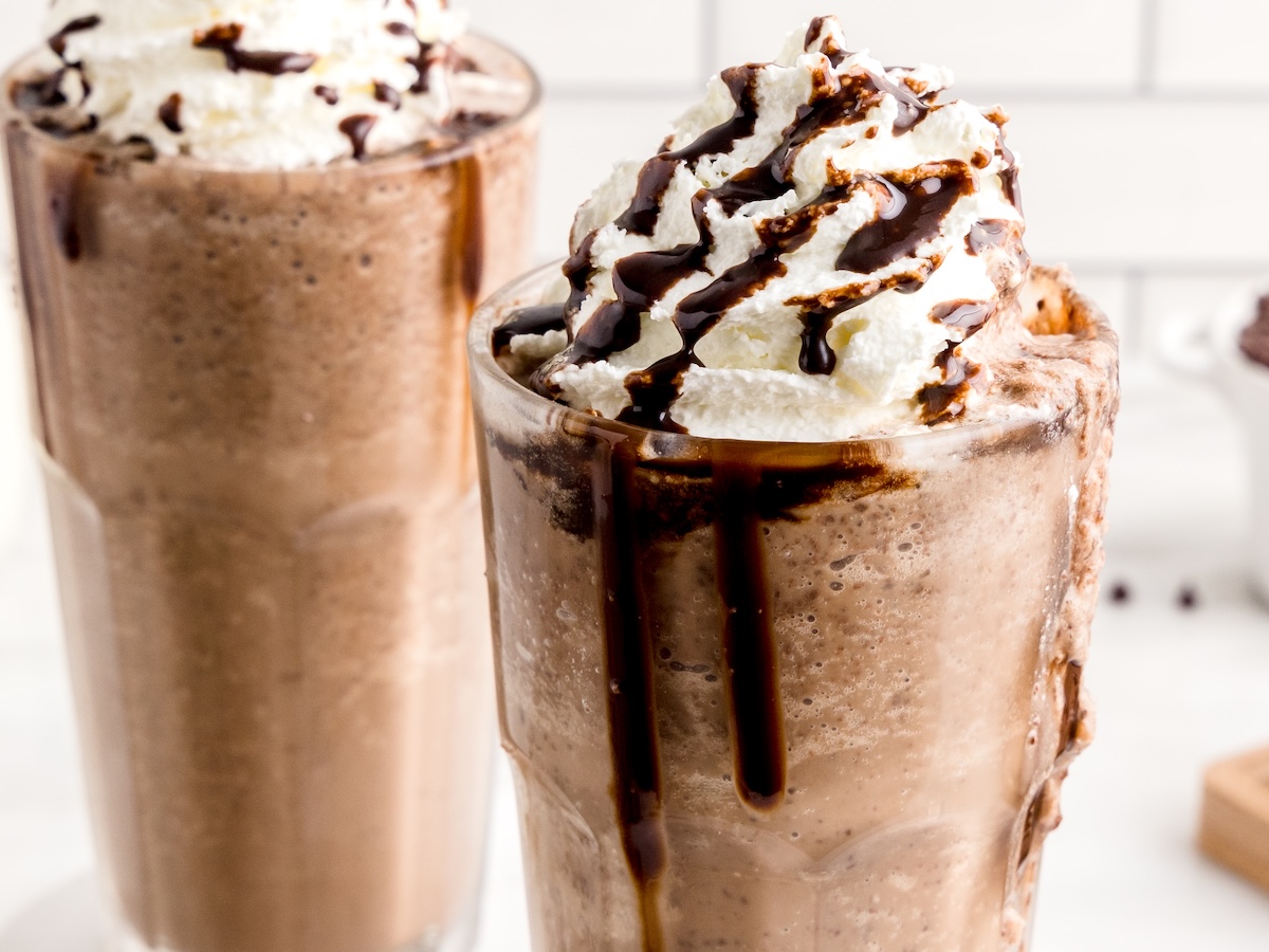 Two tall glasses filled with creamy Double Chocolate Chip Frappuccino milkshakes are topped with whipped cream and drizzled with chocolate syrup. The milkshakes look thick and cold, with chocolate dripping down the glass sides.