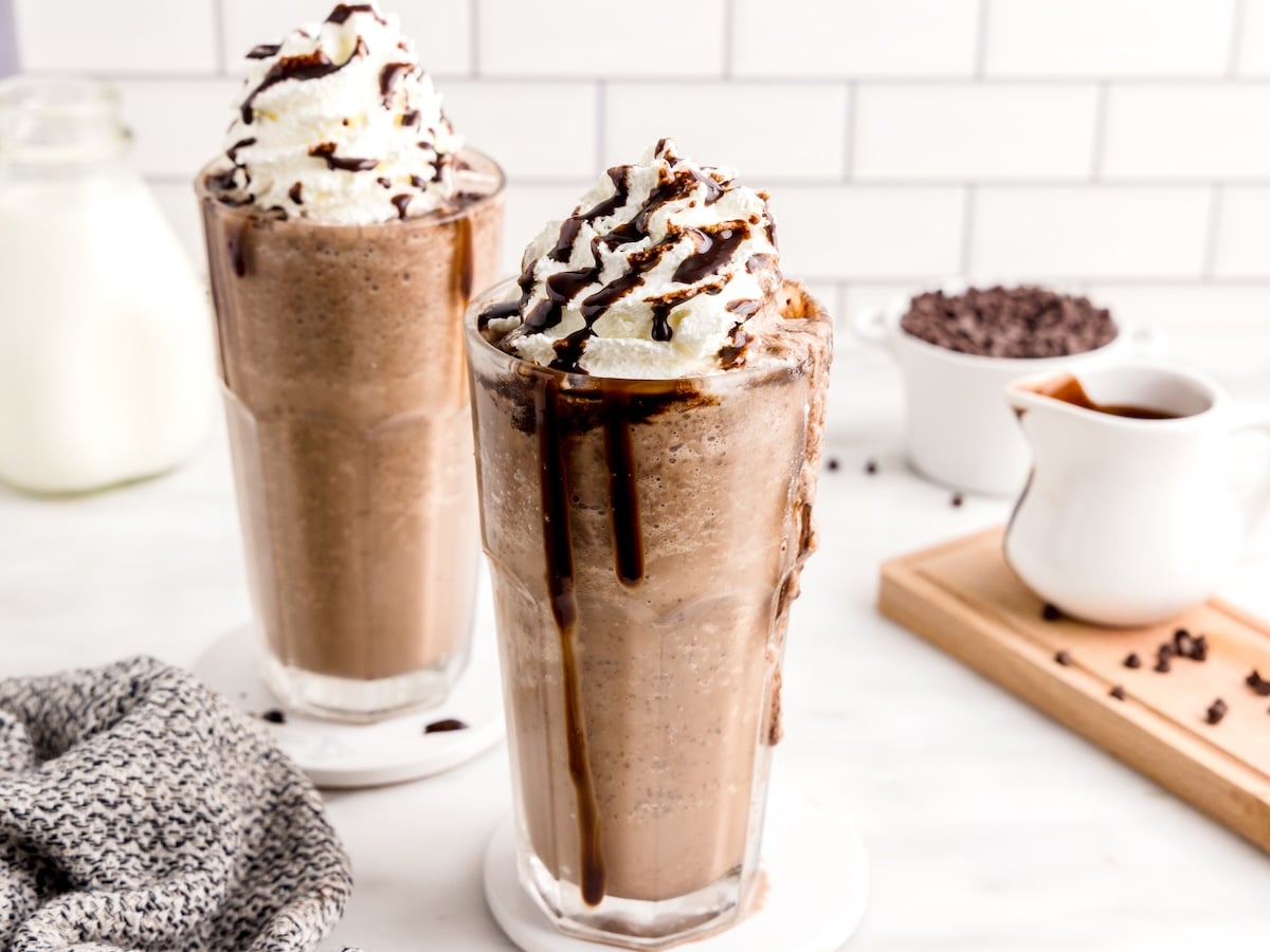 Two tall glasses of Double Chocolate Chip Frappuccino topped with whipped cream and chocolate syrup sit on coasters, with a pitcher of milk, a jug of chocolate sauce, and chocolate chips in the background.