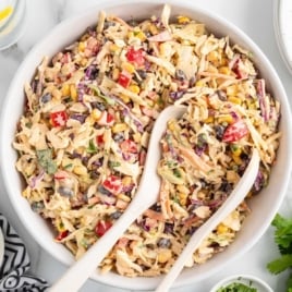 Top-down view of Mexican coleslaw in a white serving bowl with two white spoons, surrounded by fresh ingredients.