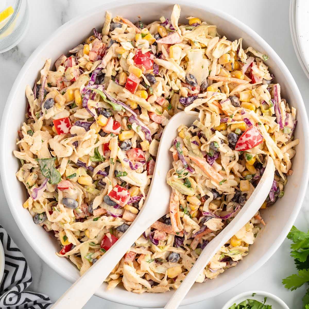 Top-down view of Mexican coleslaw in a white serving bowl with two white spoons, surrounded by fresh ingredients.