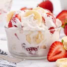 A glass bowl filled with creamy Strawberry Banana Cheesecake Salad, featuring sliced bananas and strawberries, surrounded by additional fruit slices on a white surface.