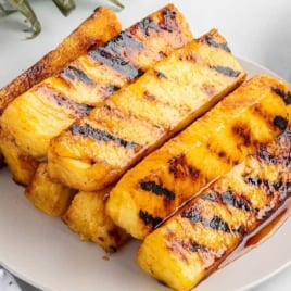 Close-up of grilled pineapple spears stacked on a white plate with caramelized edges and glistening brown sugar glaze.