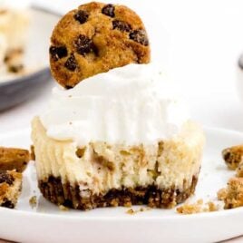 a close up shot of a Chocolate Chip Cookie Cheesecake with a bite taken out topped with a cookie on a plate