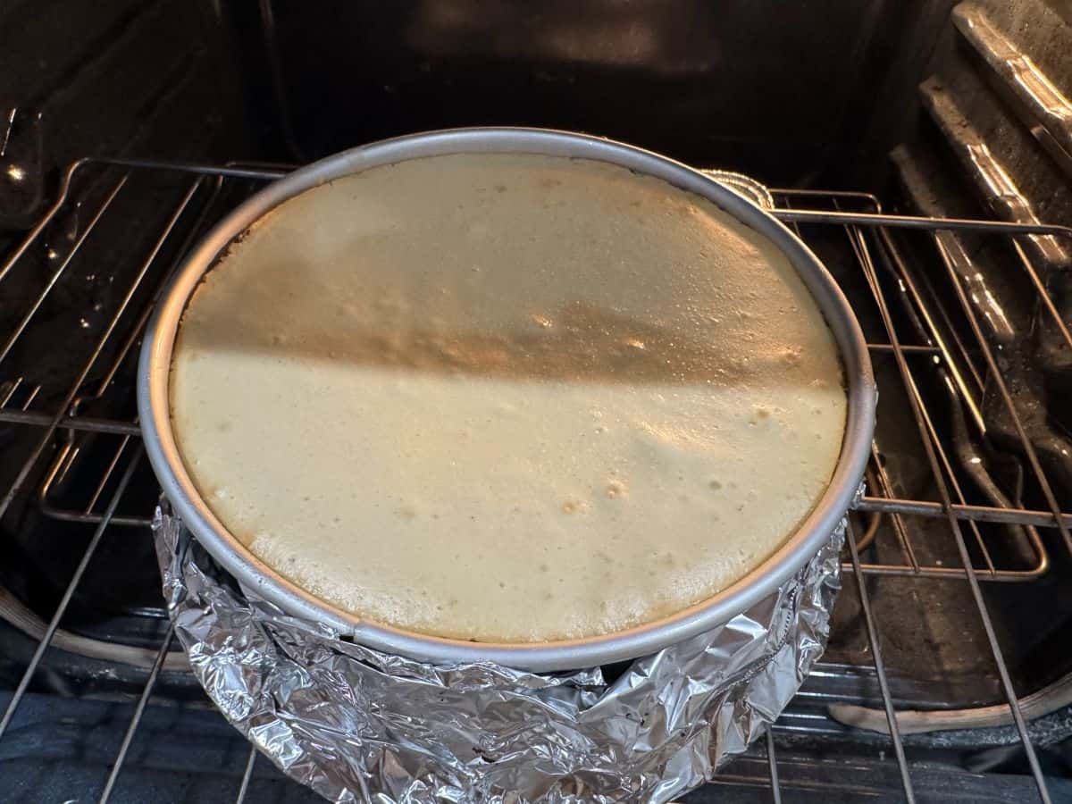 A Rolo cheesecake in a foil-wrapped springform pan baking in the oven, with a smooth, lightly golden top.