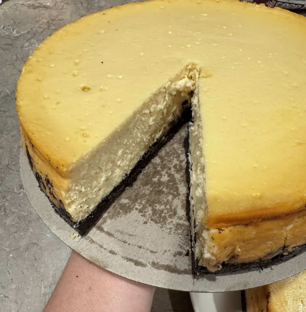 A hand holds a Rolo cheesecake with one slice cut out, showing the smooth filling over the chocolate cookie crust.