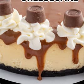 A close-up of a decadent Rolo Cheesecake, topped with caramel, whipped cream, and Rolo chocolates, with one slice cut out and served on a plate beside more Rolos and rich caramel sauce.