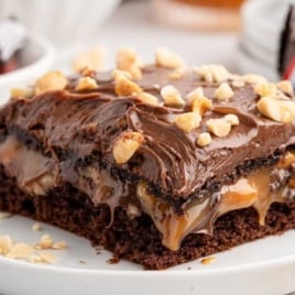 Snickers cake slice with melted caramel and chocolate frosting, garnished with chopped peanuts and Snickers bar pieces.