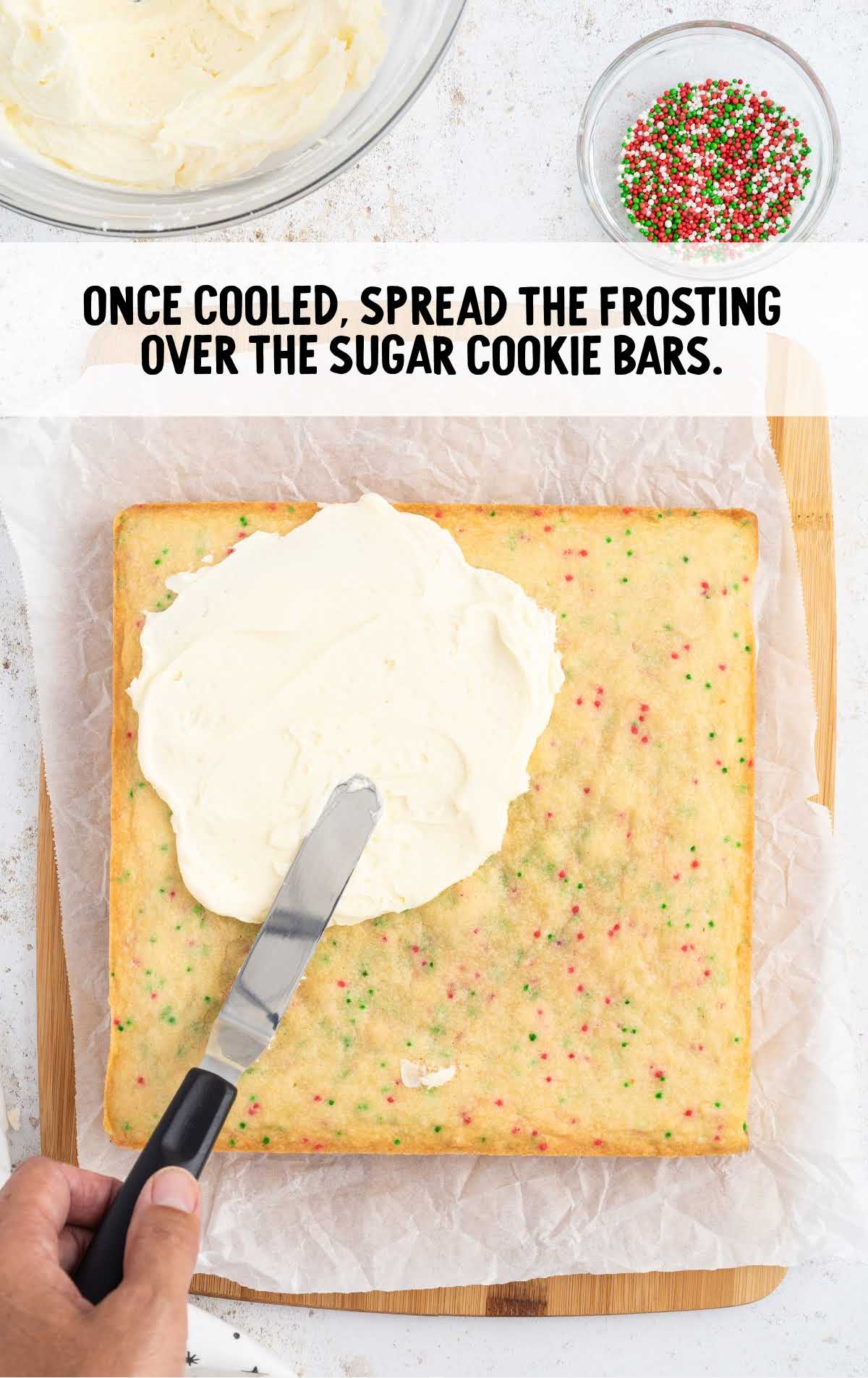 using knife spread frosting over the sugar cookie bars on a wooden board