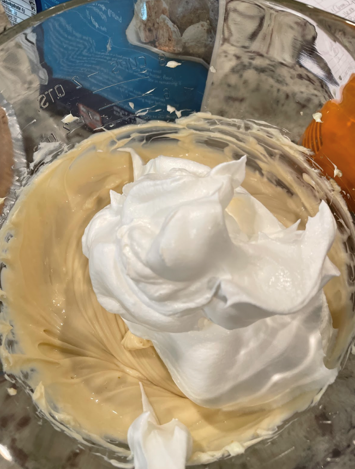 A mixing bowl contains a creamy yellow Pecan Cream Pie batter with a mound of glossy white whipped egg whites on top, ready to be folded in. Baking ingredients and tools are visible in the background.