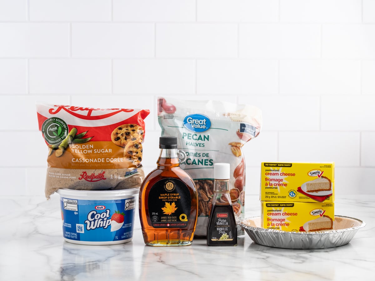 A variety of baking ingredients on a marble counter&mdash;perfect for Pecan Cream Pie&mdash;including brown sugar, pecan halves, maple syrup, vanilla extract, cream cheese, whipped topping, and a graham pie crust against a white tile background.