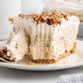 A close-up of a slice of Pecan Cream Pie topped with chopped pecans on a white plate, with a fork cutting into the pie. The creamy filling appears fluffy and light, and the golden crust looks perfectly crumbly.