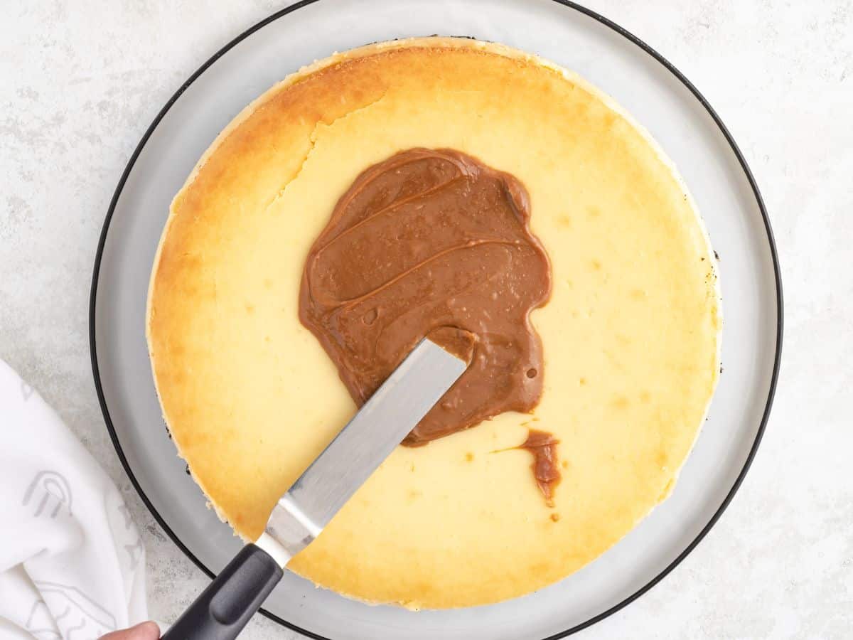 An offset spatula spreads dulce de leche over the top of a plain Rolo cheesecake on a glass plate.