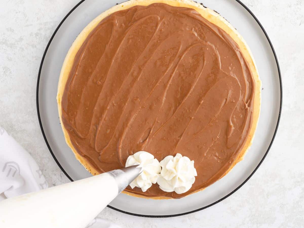 Whipped cream being piped in swirls around the edge of a Rolo cheesecake topped with dulce de leche.