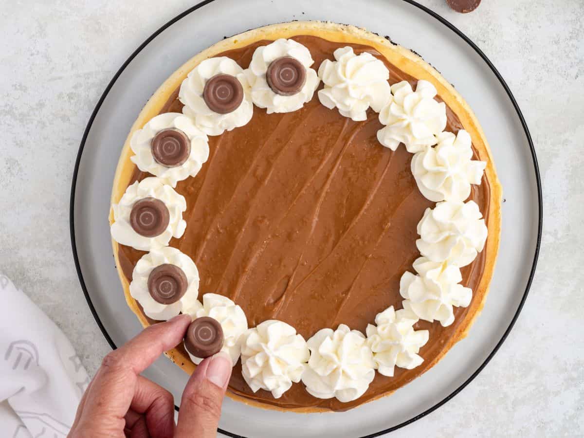 A hand places a Rolo on a whipped cream swirl on a Rolo cheesecake topped with dulce de leche.