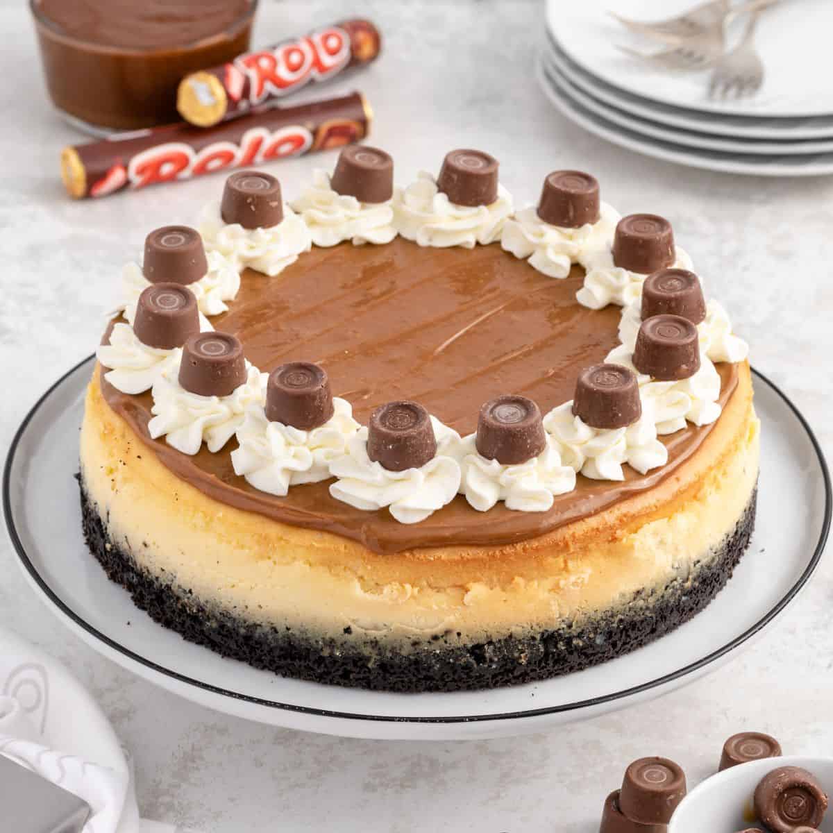 Rolo Cheesecake with a chocolate cookie crust, topped with caramel, whipped cream, and Rolo chocolates, displayed on a white plate. Extra Rolos and dessert plates are in the background.