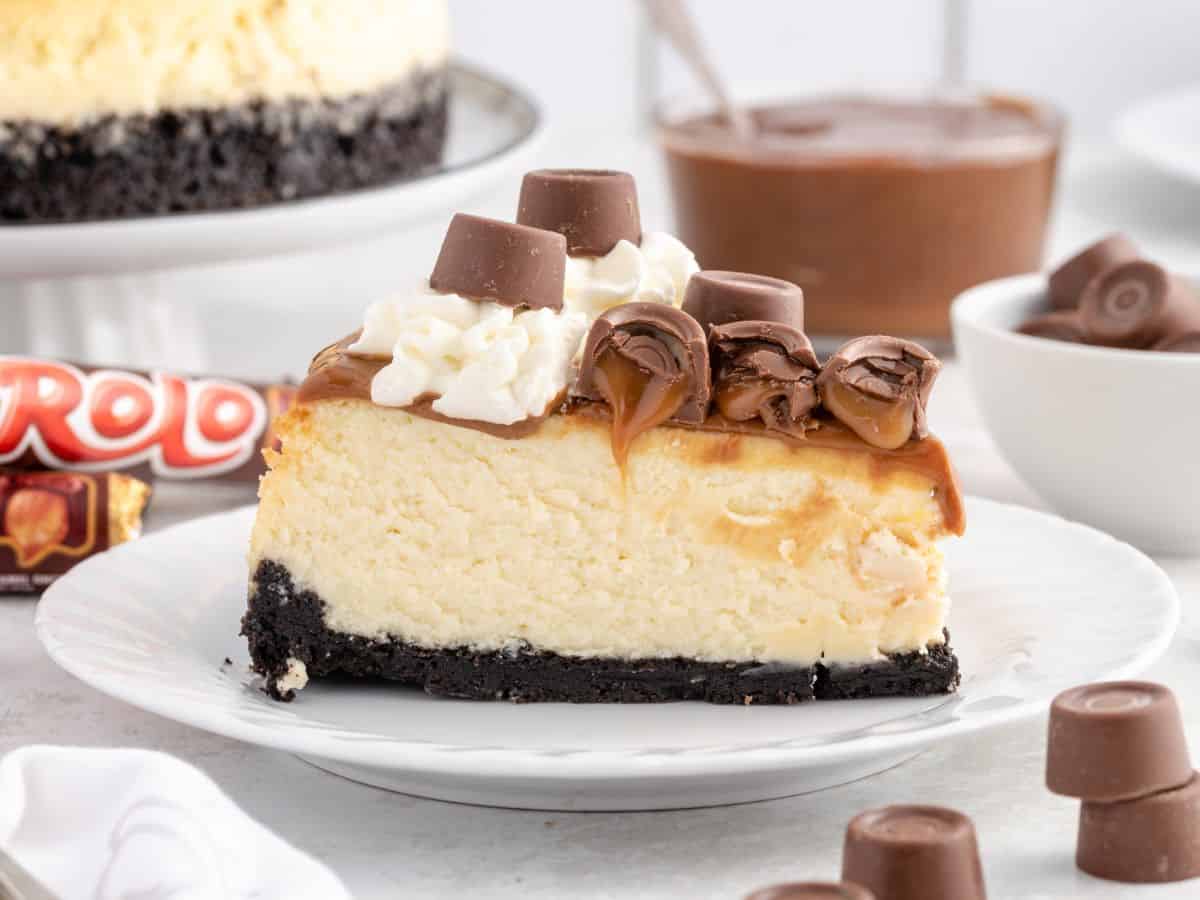 A slice of Rolo cheesecake on a white plate, with a chocolate cookie crust, dulce de leche over the top, a swirl of whipped cream and a Rolo, with more Rolos in the background.