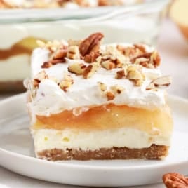 A slice of Apple Delight layered dessert with a crumbly crust, creamy filling, apple topping, whipped cream, and chopped pecans sits on a white plate. The rest of the Apple Delight can be seen in the background.