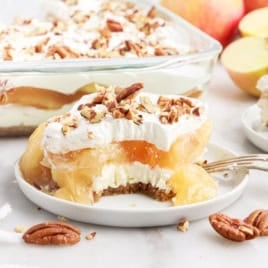 A plate with layered Apple Delight dessert topped with whipped cream and chopped pecans; a bite is missing. More dessert, apples, and pecans are in the background.