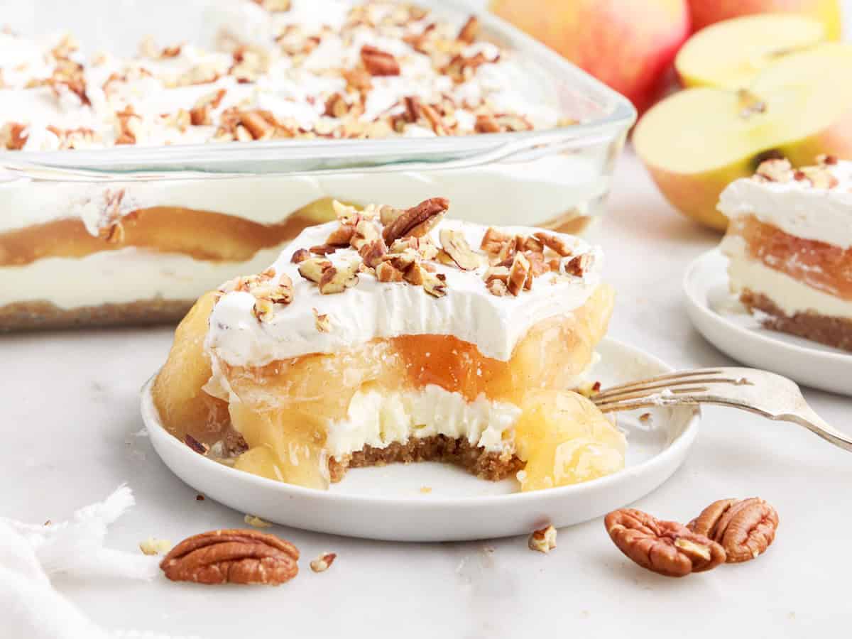 A plate with layered Apple Delight dessert topped with whipped cream and chopped pecans; a bite is missing. More dessert, apples, and pecans are in the background.