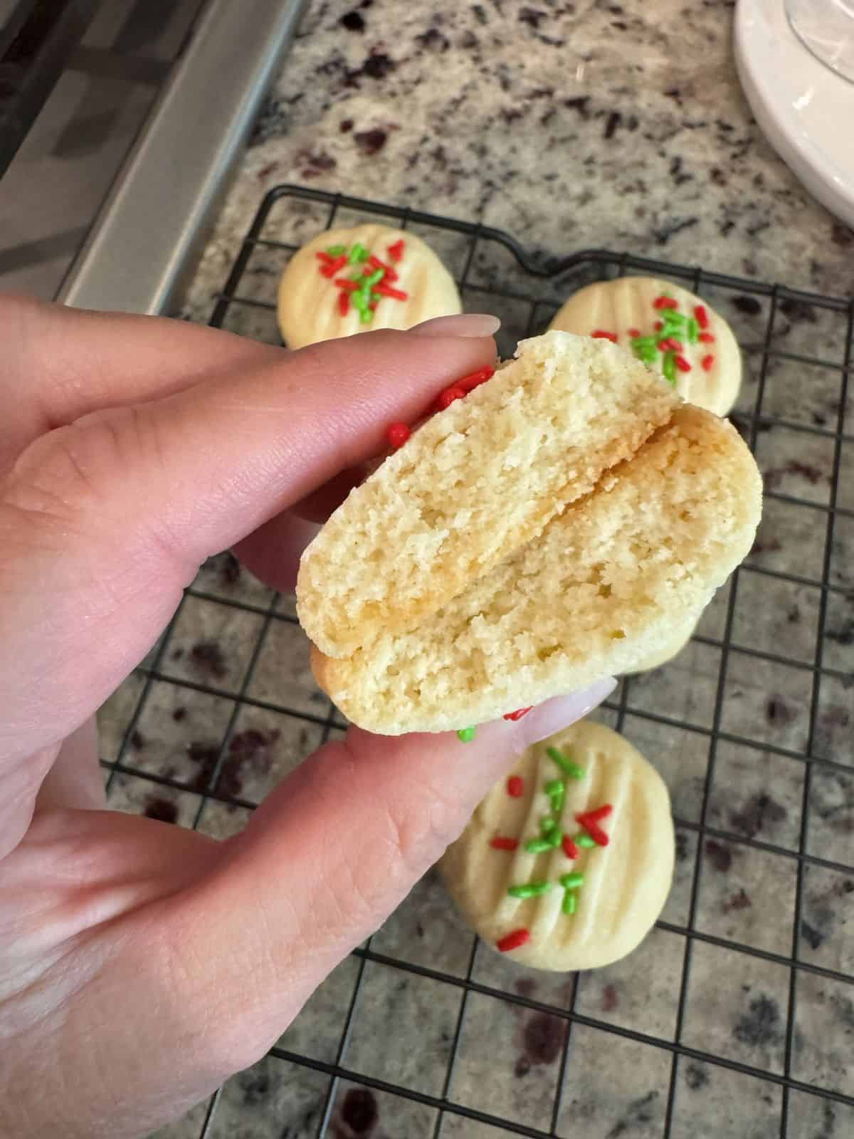 A hand holds a half-eaten Christmas Shortbread Cookie over a cooling rack with more cookies topped with red and green sprinkles on a granite countertop.