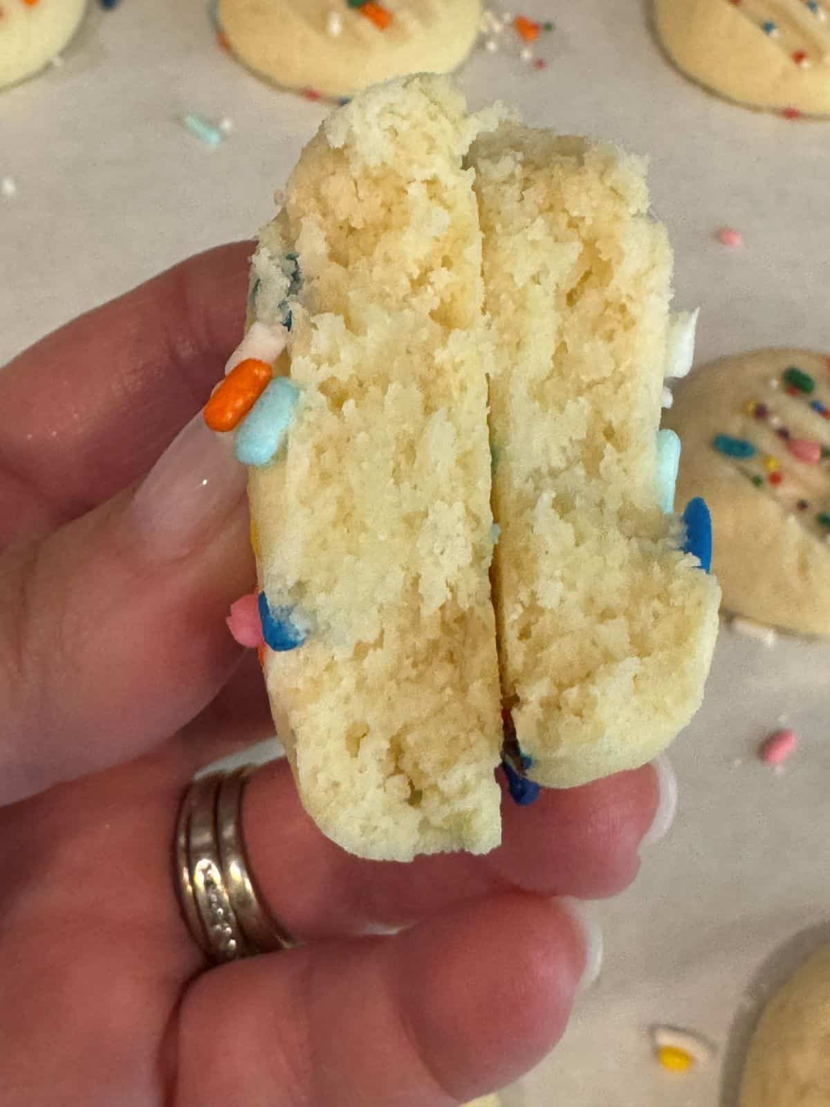 A hand holds a soft, sugar cookie broken in half, revealing a fluffy interior reminiscent of classic Christmas Shortbread Cookies. The cookie is decorated with colorful sprinkles, while other cookies are visible in the background.