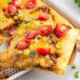 a close up shot of Taco Pizza in a baking pan with a piece taken out with a wooden spatula