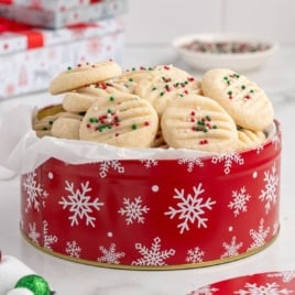 A red tin with white snowflakes holds a pile of Christmas Shortbread Cookies, each round cookie topped with festive red, green, and white sprinkles. Some wrapped gifts and a bowl of sprinkles are in the background on a white surface.