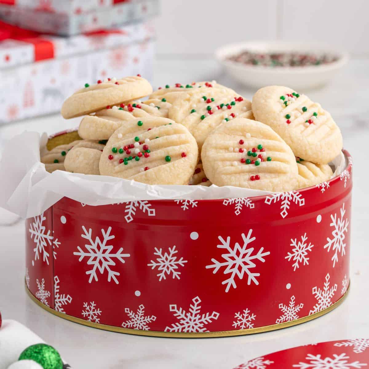 A red tin with white snowflakes holds a pile of Christmas Shortbread Cookies, each round cookie topped with festive red, green, and white sprinkles. Some wrapped gifts and a bowl of sprinkles are in the background on a white surface.