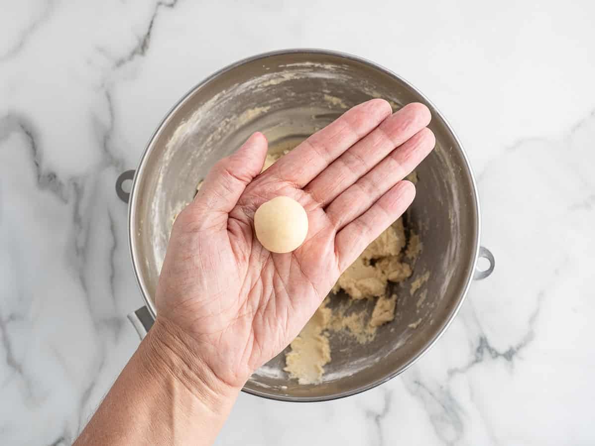 A hand holds a small ball of Christmas Shortbread Cookies dough over a metal mixing bowl filled with more dough, on a white marble surface.